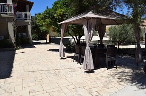 An outdoor patio with a covered seating area, stone flooring, and surrounding greenery. - Villa Miranda (Zakynthos)