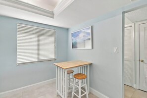A hallway with a light blue wall, a window with blinds, a framed picture, a white door with louvered glass, and a white bar stool with a wooden top. - Loggerhead Cay 562 - Partial gulf view, beach side vacation rental condo (Sanibel)