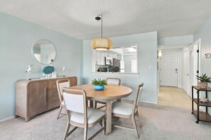 A dining area with a round table, chairs, a wooden cabinet, a mirror, and a hanging light fixture. - Loggerhead Cay 562 - Partial gulf view, beach side vacation rental condo (Sanibel)