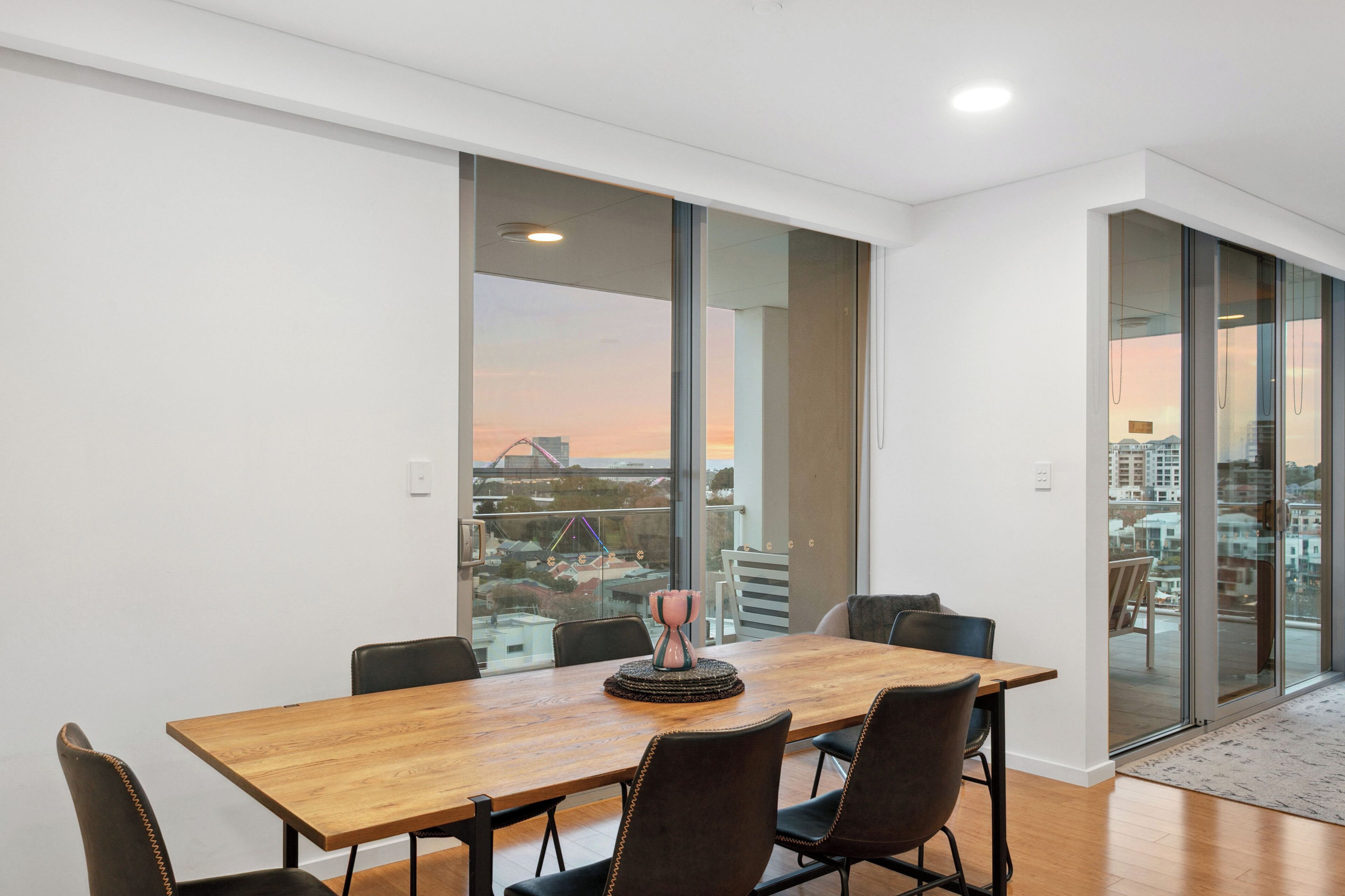 A modern dining area with a wooden table and chairs, large windows with a city view, and a glass door leading to an outdoor balcony.