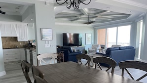 A modern kitchen with white cabinets, a dining area with a wooden table and chairs, a living room with blue sofas and a flat-screen TV, and a ceiling fan.