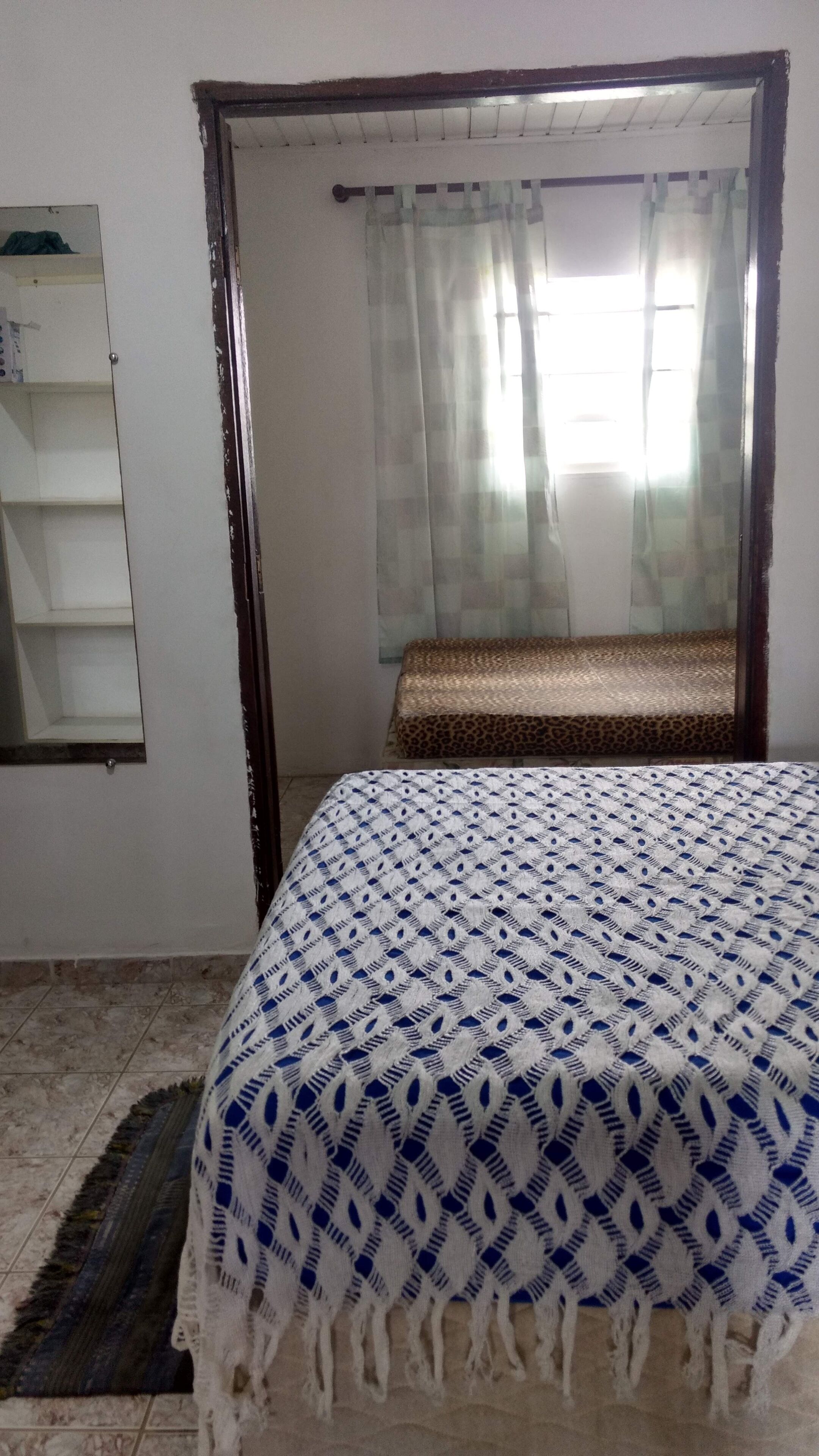 Un dormitorio con cama, un espejo, una ventana con cortinas y un pequeño banco.