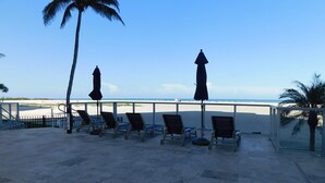 A beachside patio with lounge chairs and umbrellas, overlooking a sandy beach and the ocean.