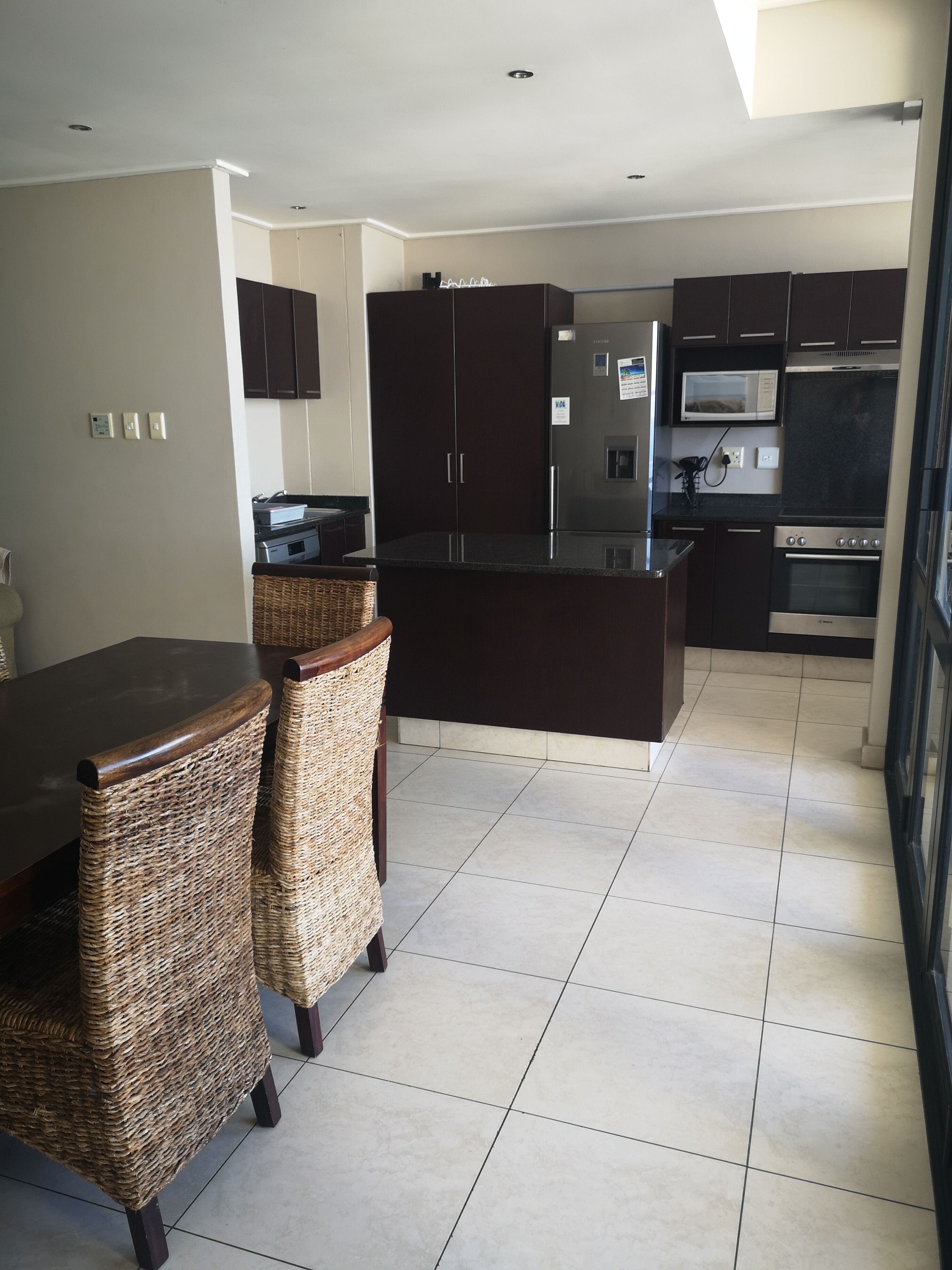 A modern kitchen with dark cabinetry, a stainless steel refrigerator, a microwave, and an oven. There is a dining area with a dark table and wicker chairs.