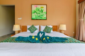 A hotel room with a bed, two pillows, two bedside lamps, a painting of lotus flowers, and a towel arranged in the shape of a swan on the bed.