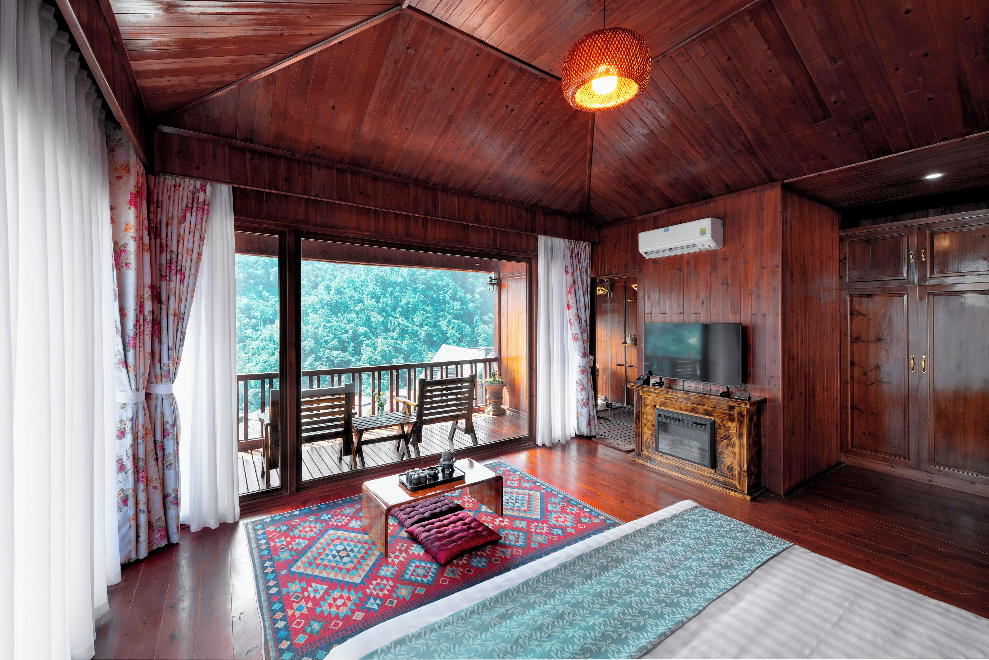 A wooden-floored living room with a television, a fireplace, and a balcony with a view of trees.