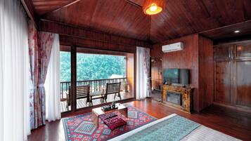 A wooden-floored living room with a television, a fireplace, and a balcony with a view of trees.