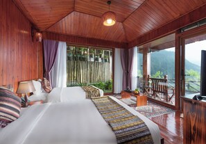 A wooden cabin room with a bed, a table, a chair, and a view of the outdoors.