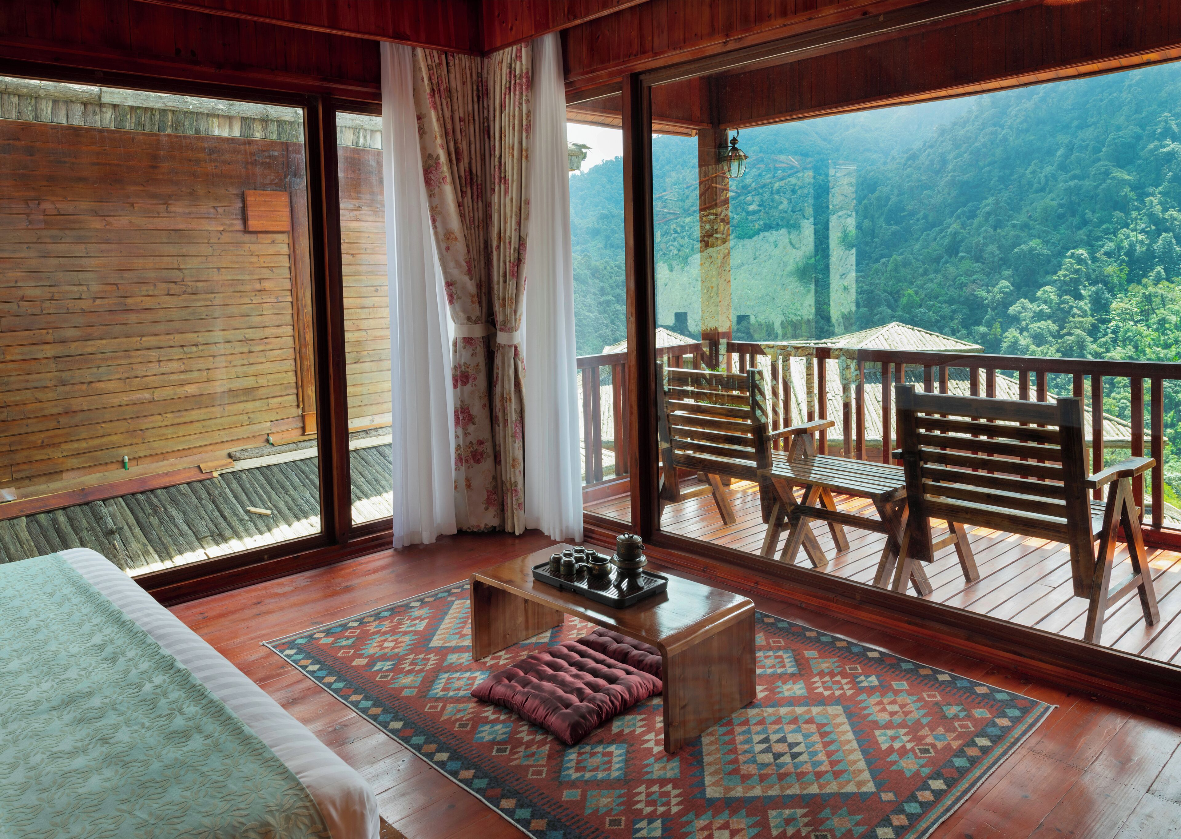 A wooden-floored room with a large window, a wooden bench, a small table, and a patterned rug.