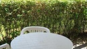Mesa redonda blanca con cuatro sillas en un entorno exterior con un sendero pavimentado y setos verdes.