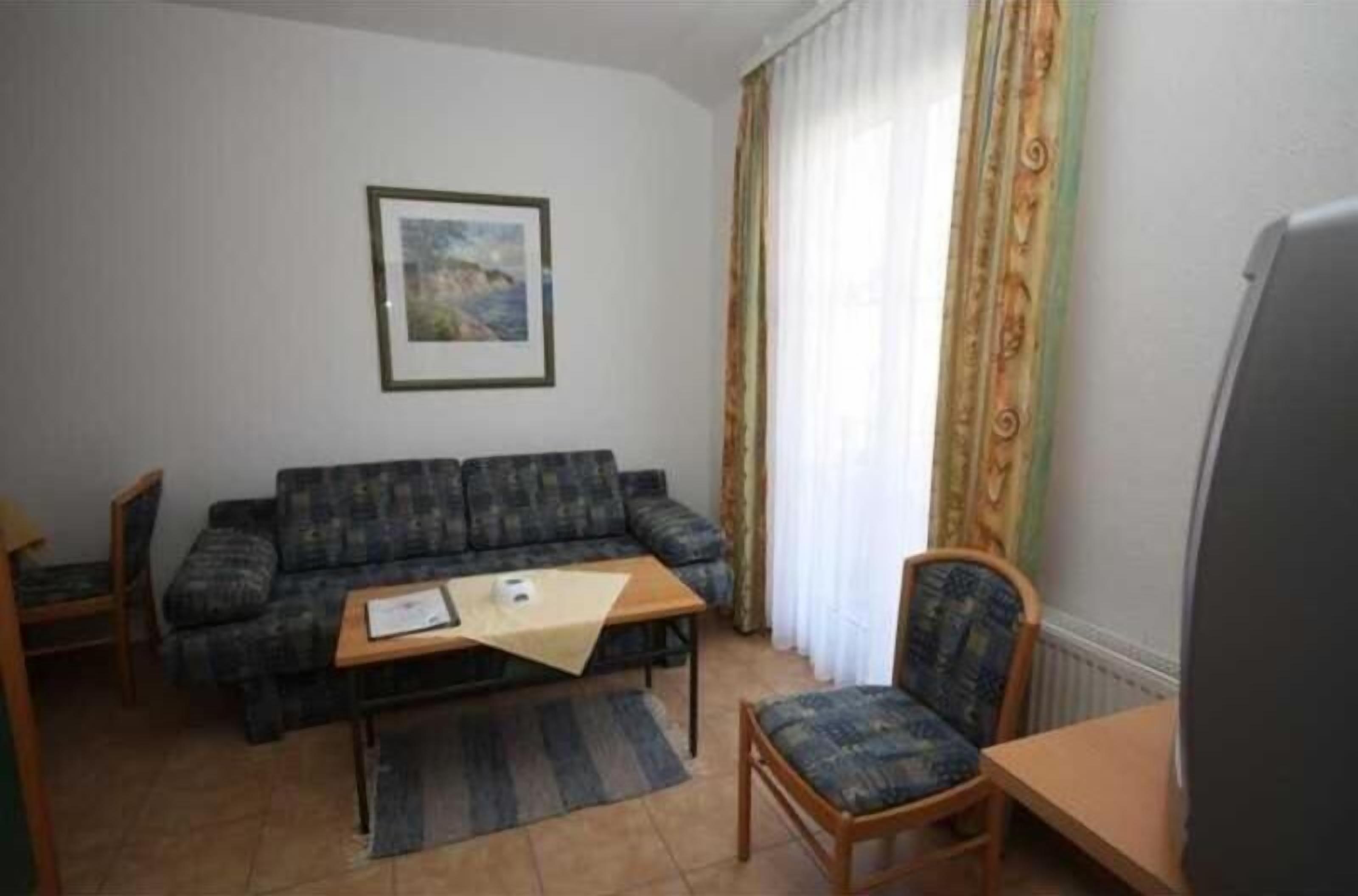 A living room with a sofa, a coffee table, and two chairs. There is a framed picture on the wall and a window with curtains.