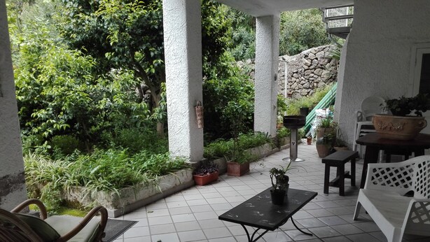 Zona de terrassa coberta amb paviment de rajola, una cadira de vimet, una taula petita i plantes en testos.