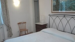 Un dormitori amb un llit, una cadira, una tauleta de nit, una làmpada i un quadre a la paret.