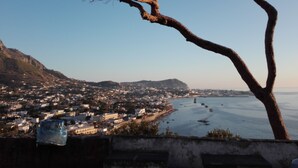 Un poble costaner amb un arbre destacat al primer pla, vista clara del mar i una serralada de muntanyes a la distància.