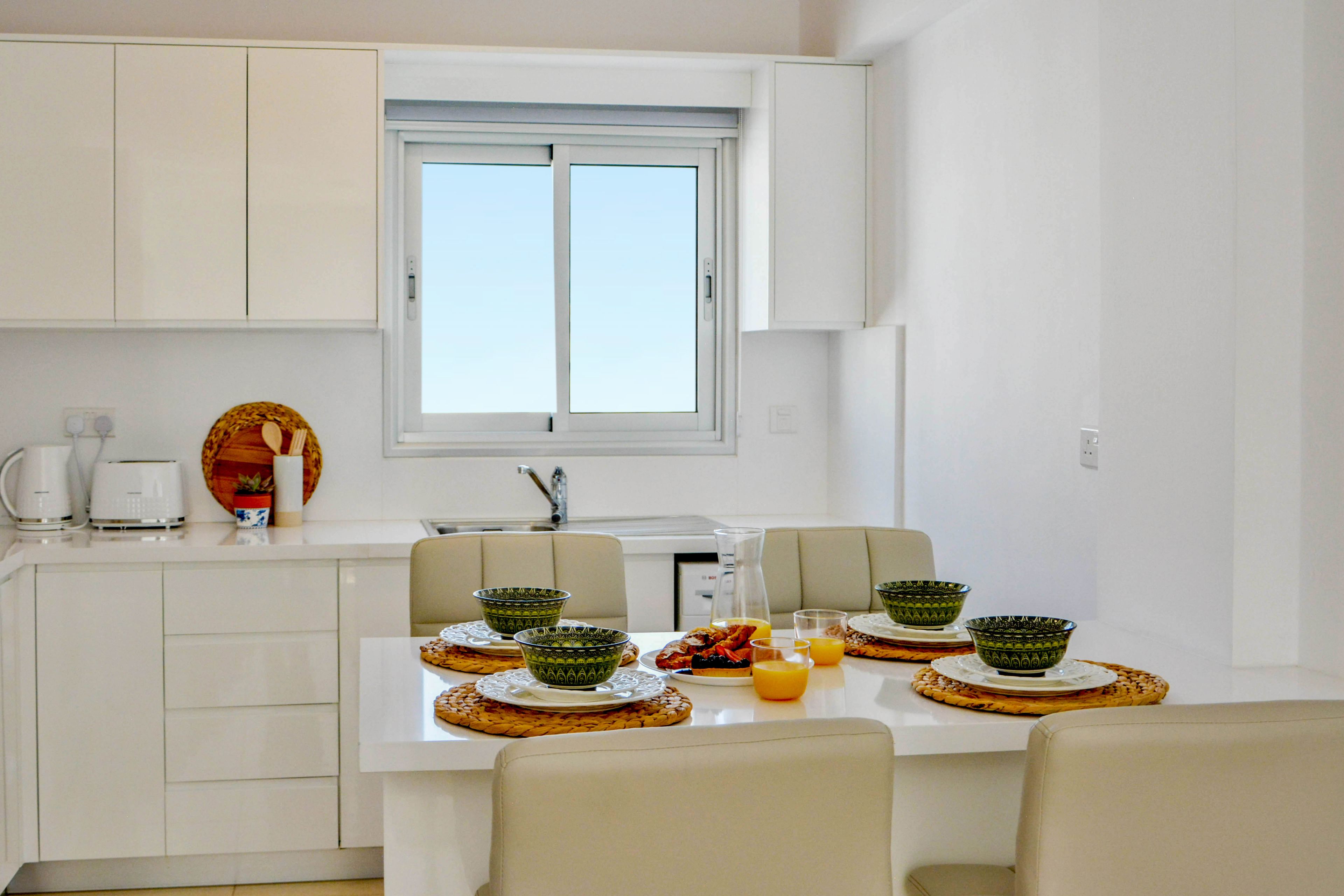 A modern kitchen with white cabinets, a dining table set with green bowls, plates, and a pitcher of orange juice.