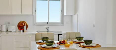A modern kitchen with white cabinets, a dining table set with green bowls, plates, and a pitcher of orange juice.
