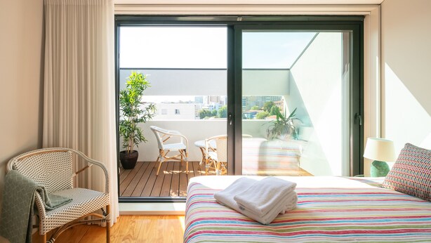 A hotel room with a bed, a chair, a table, a balcony with a plant, and a view of the city. - Porto Insight Apartment Cedofeita- Balcony & Parking (Porto)