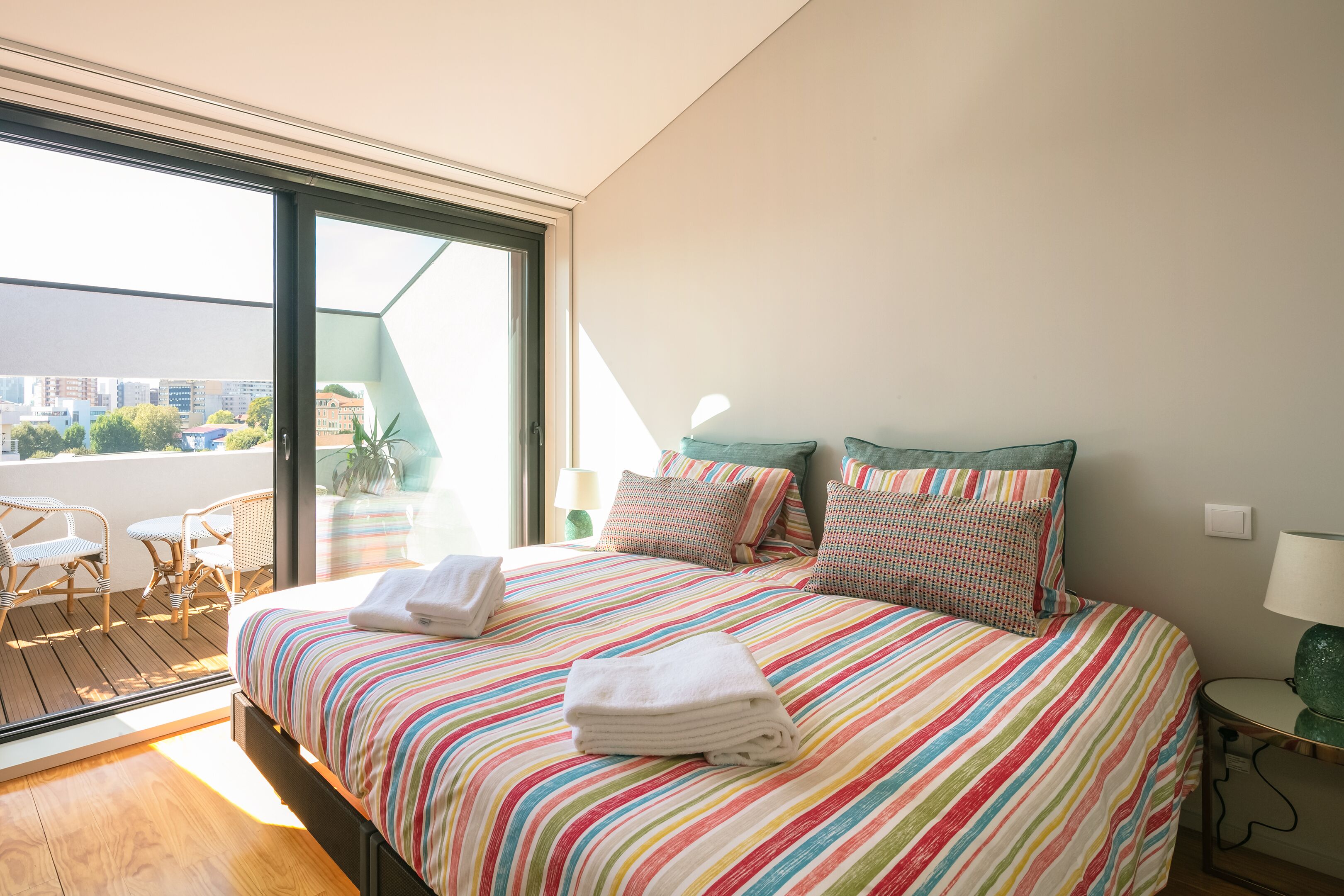 A bedroom with a large bed, two pillows, a bedside table with a lamp, and a view of the city through a large window.
