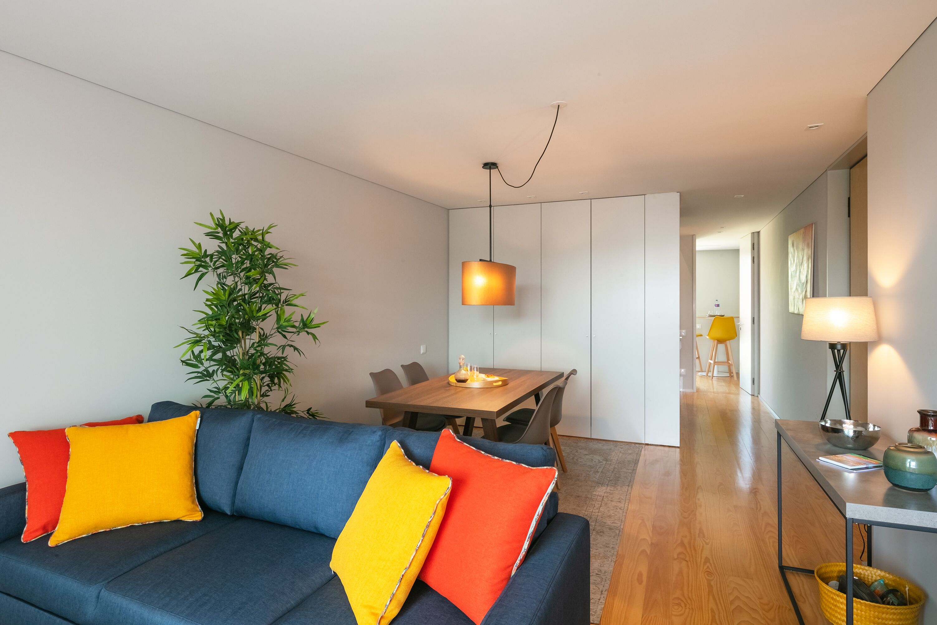 Ein modernes Wohnzimmer mit einem dunkelblauen Sofa, leuchtend orangefarbenen und roten Kissen, einem Essbereich mit einem Holztisch und Stühlen sowie einem kleinen Beistelltisch mit einer Lampe.
