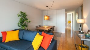 A modern living room with a dark blue sofa, bright orange and red cushions, a dining area with a wooden table and chairs, and a small side table with a lamp. - Porto Insight Apartment Cedofeita- Balcony & Parking (Porto)
