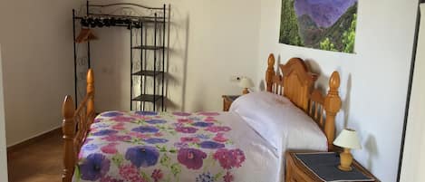 Un dormitori amb un llit de fusta, un llençol floral, una prestatgeria de metall, una tauleta de nit de vimet i una pintura de paisatge de muntanya.