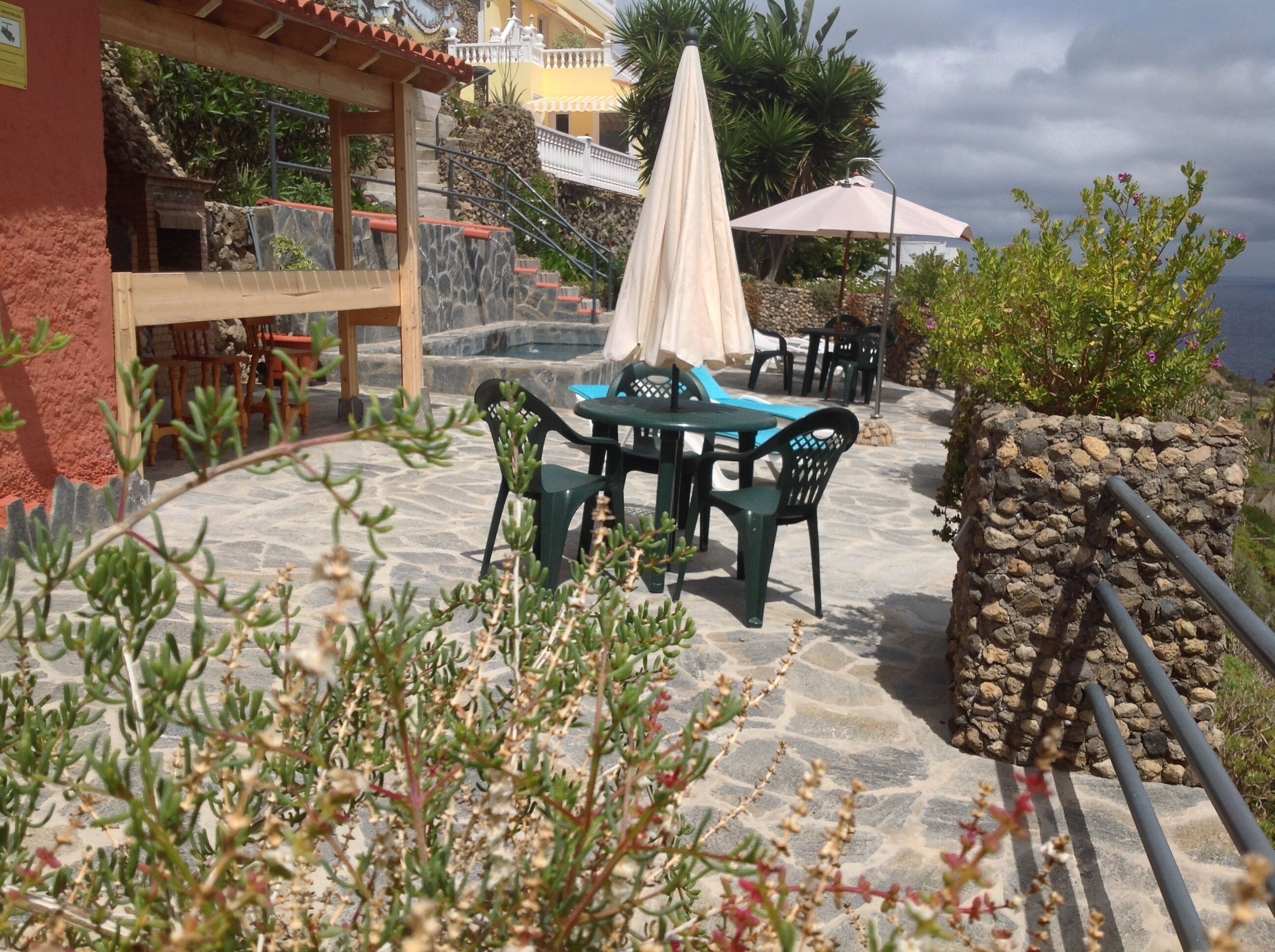 An outdoor patio area with stone flooring, green plastic chairs, a white umbrella, and a stone wall.