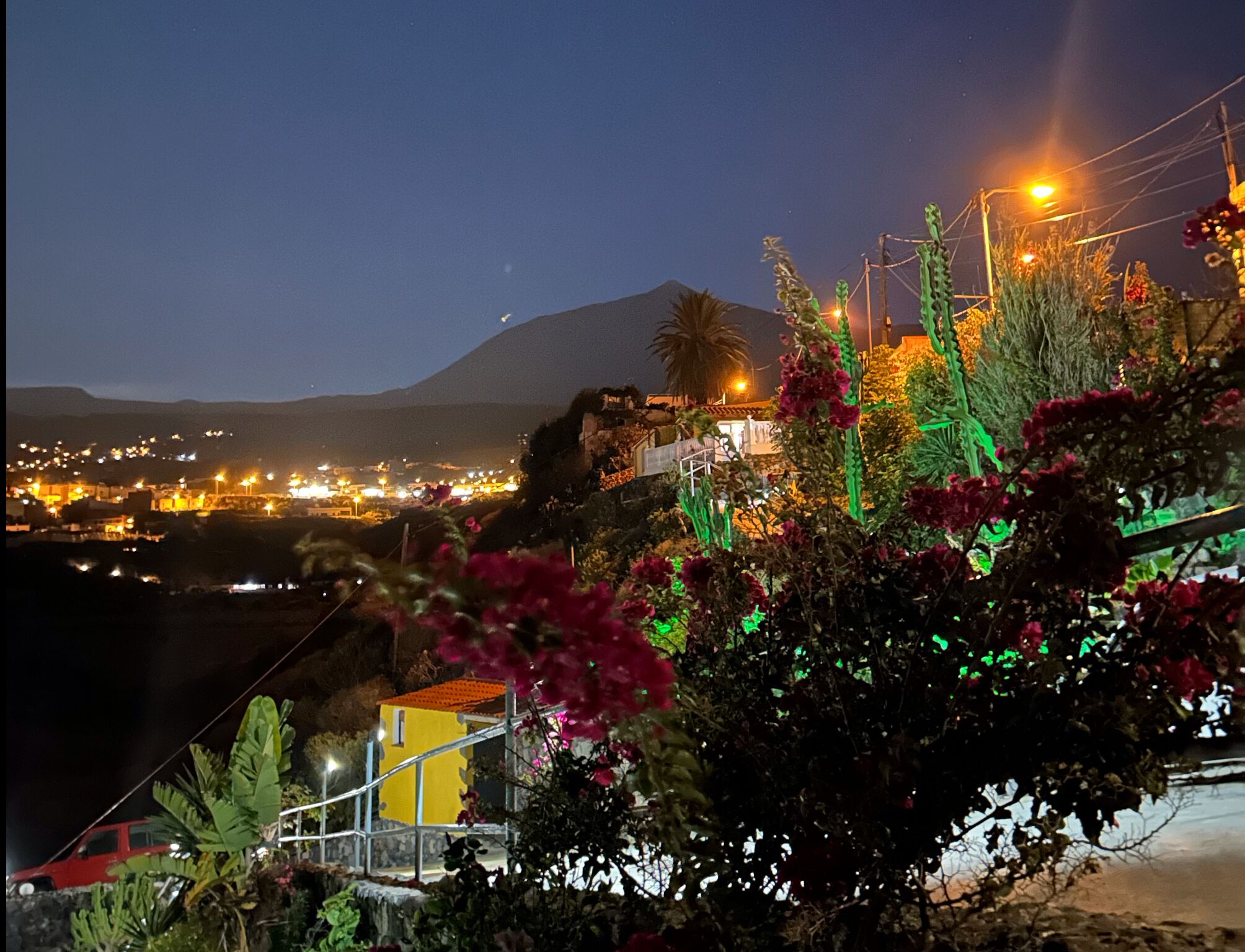 Une vue nocturne d’une ville, avec une montagne en arrière-plan, un bâtiment jaune illuminé et des structures éclairées en vert.