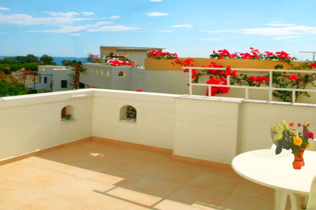 Una terraza en la azotea con una mesa y sillas blancas, flores rojas y un cielo azul despejado.