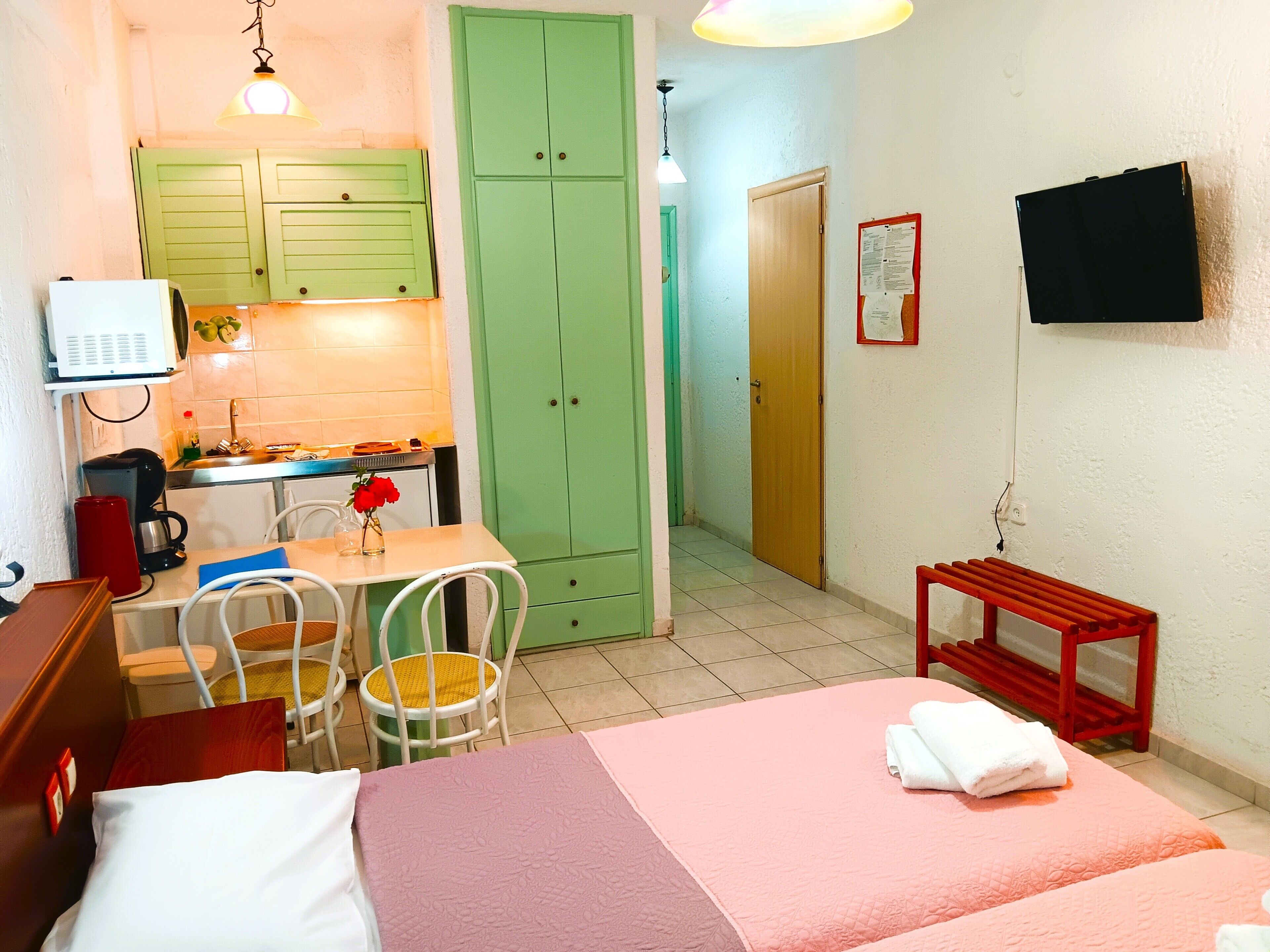 A compact room with a bed, a desk with a chair, a small table with a red bench, a green door, and a TV mounted on the wall.
