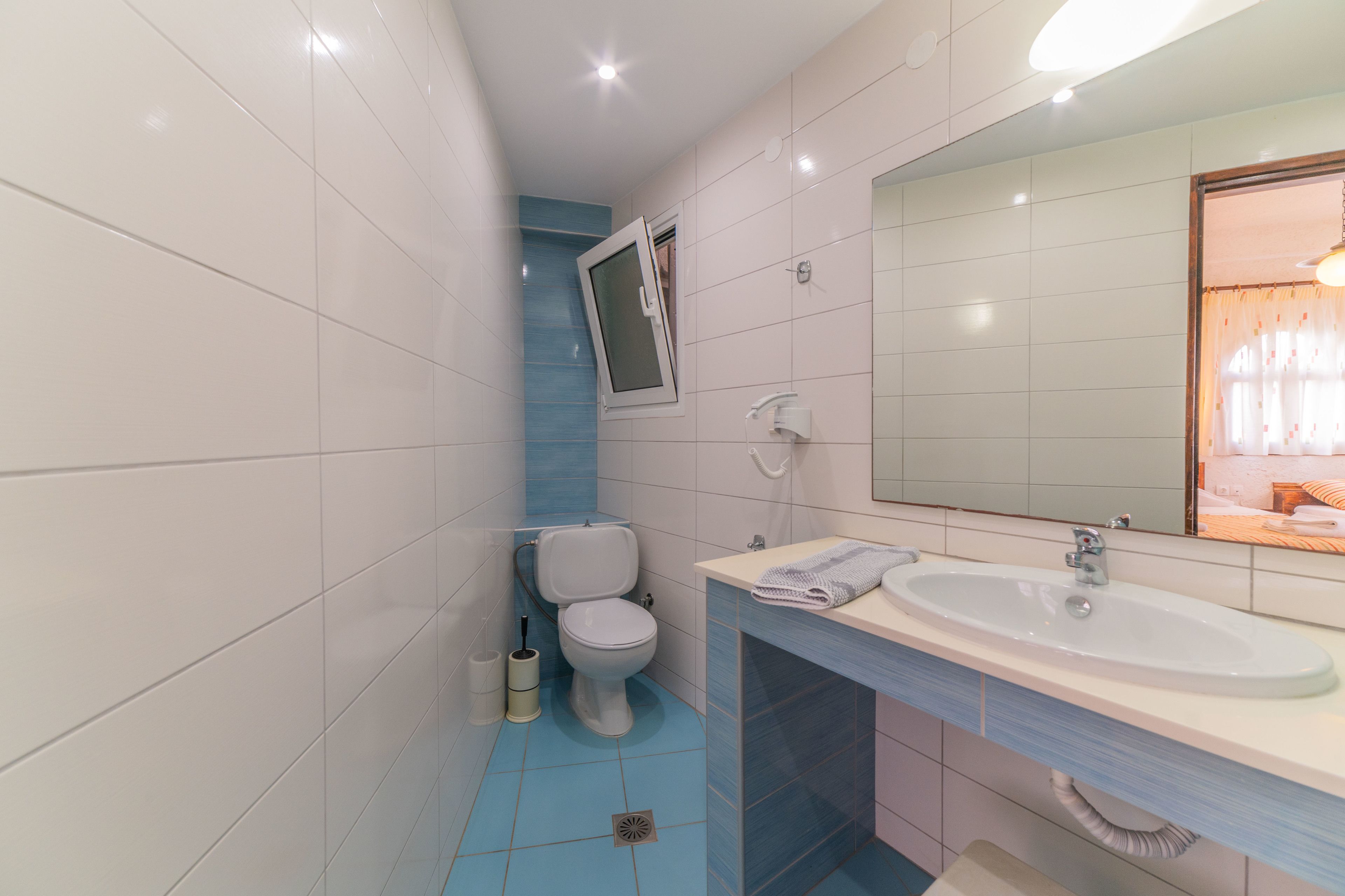Un baño con azulejos blancos y azules, un inodoro blanco, un lavamanos con espejo y una ventana con cortinas.