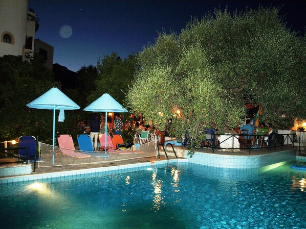 A swimming pool with lounge chairs and umbrellas, surrounded by trees and a building. - Kastro Studios Apartments (Ierapetra)