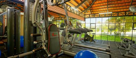A gym with exercise equipment, a treadmill, and a view of greenery through large windows.