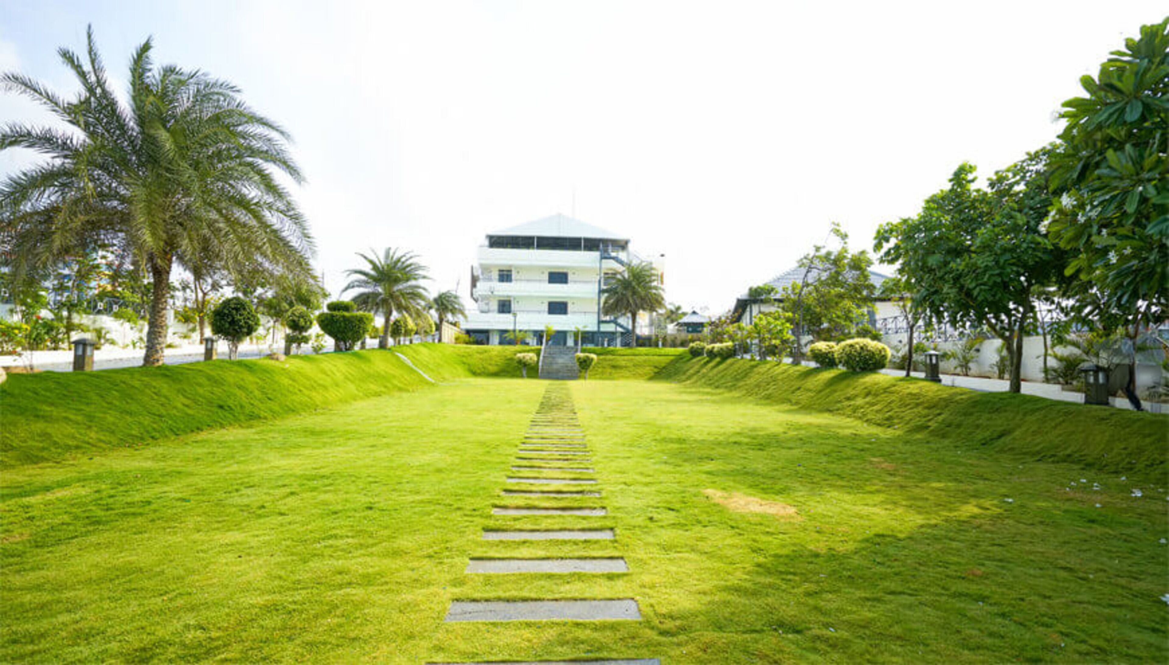 A well-maintained grassy pathway leading to a modern building with multiple floors, surrounded by lush greenery and palm trees.