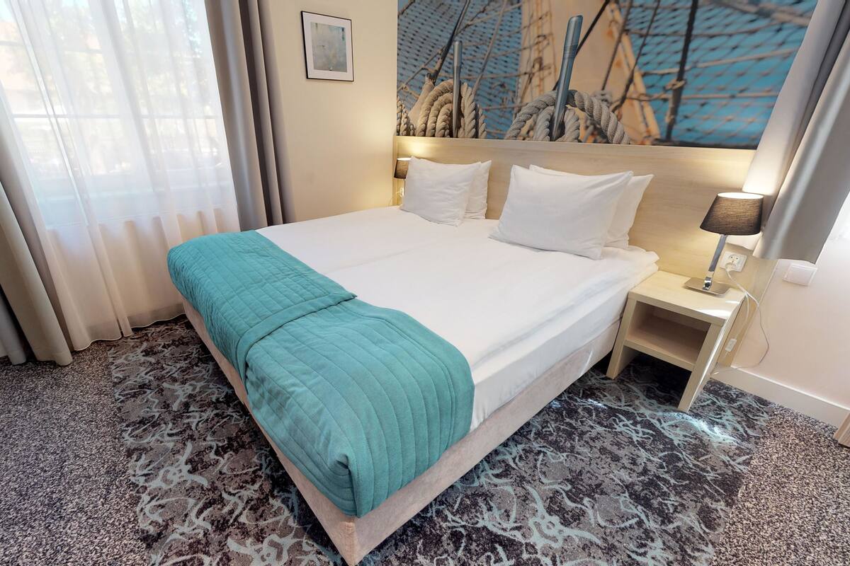 A hotel room with a large bed, a headboard with a painting, a nightstand with a lamp, and a window with curtains.