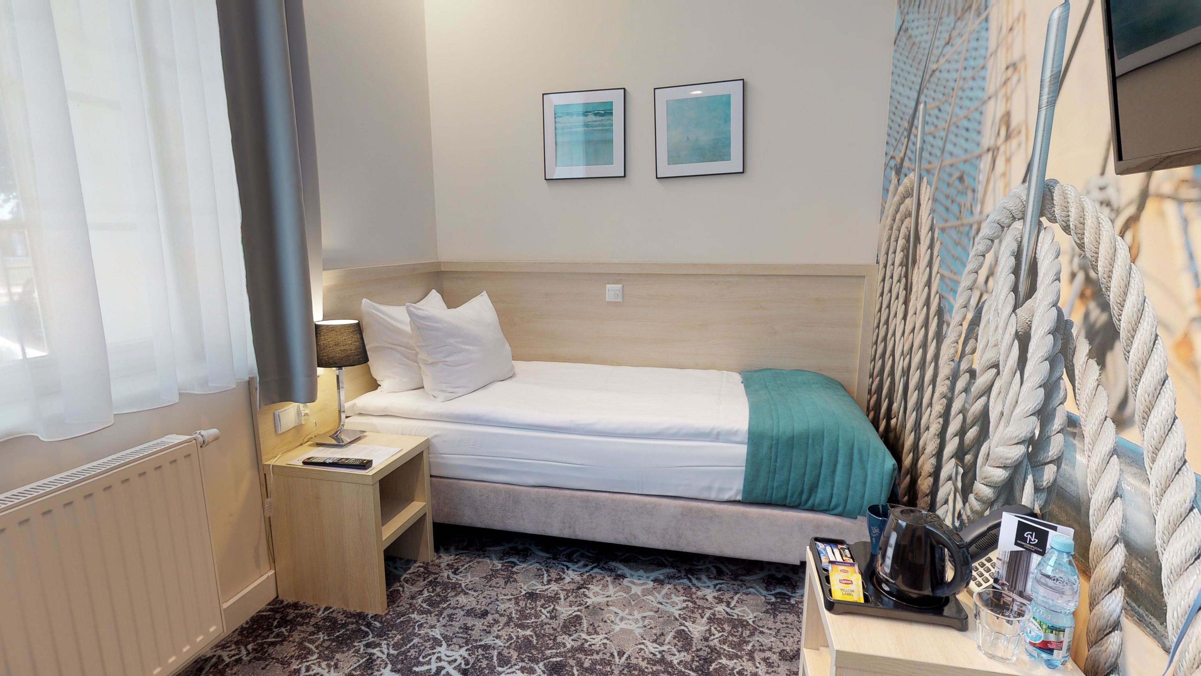 Une chambre d’hôtel comprenant un lit, une table de chevet, une lampe, un tableau encadré, une fenêtre avec des rideaux et un téléviseur fixé au mur.