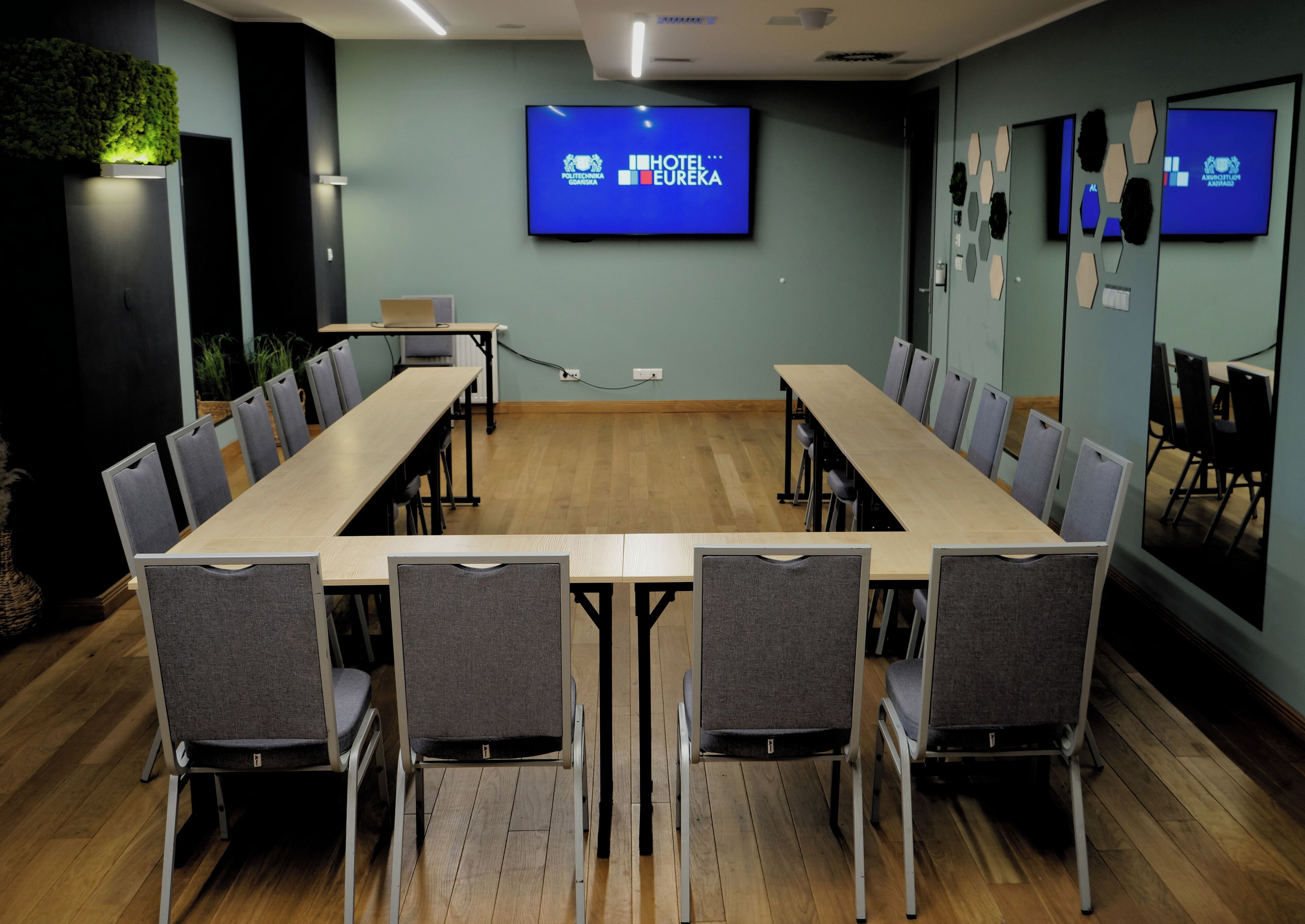 Een vergaderruimte met een lange tafel en stoelen, een televisiescherm waarop een logo wordt getoond, en een plant aan de muur.
