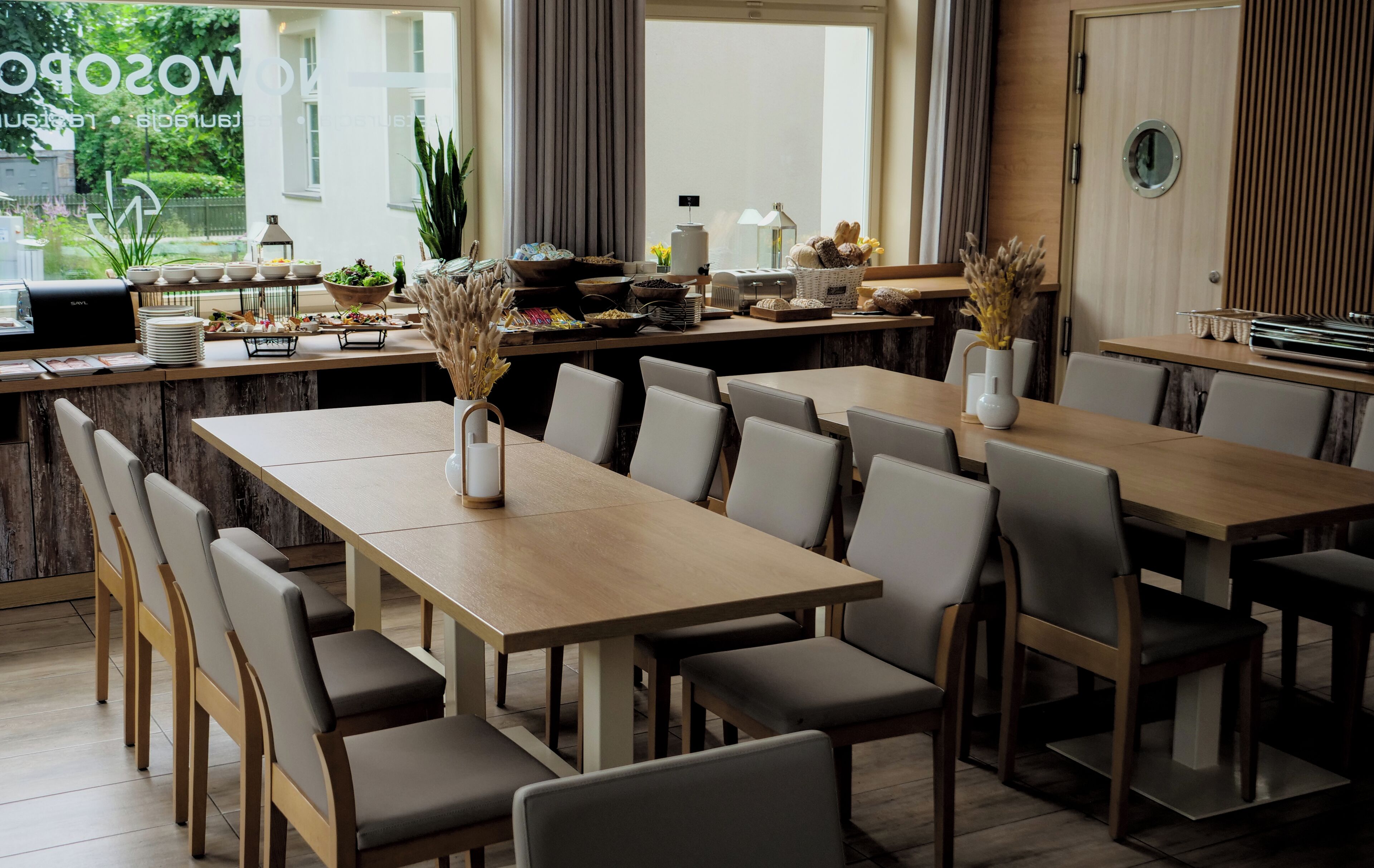 A modern dining area with long tables and chairs, a buffet setup, and a view of greenery outside.