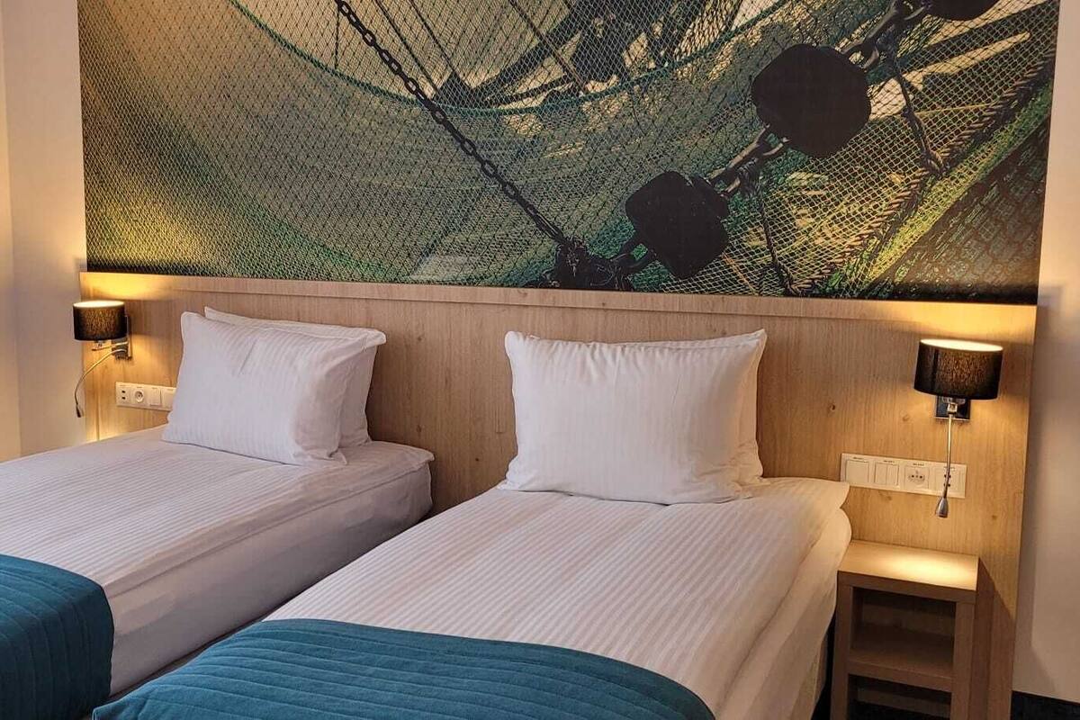 A hotel room with two single beds, a large window with sheer curtains, and a wall-mounted artwork.