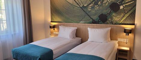 A hotel room with two single beds, a large window with sheer curtains, and a wall-mounted artwork.