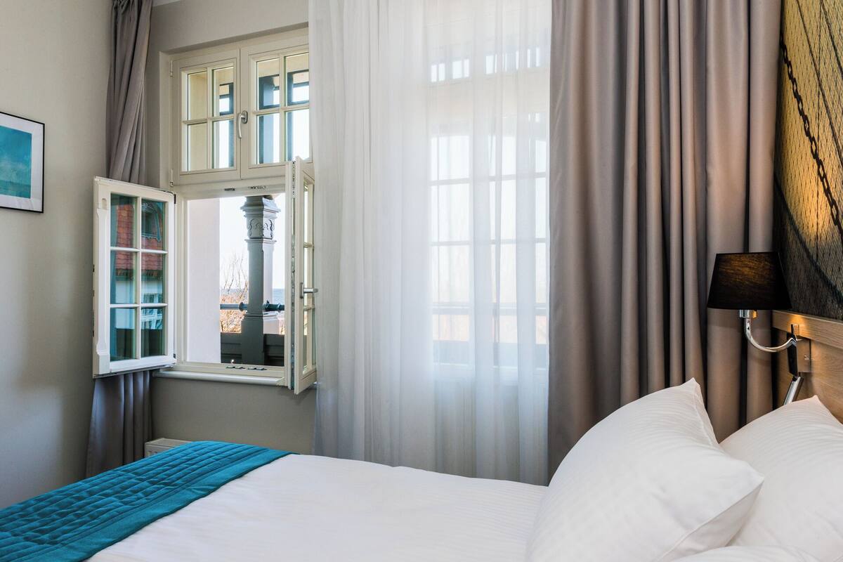A hotel room with a bed, a window with curtains, a lamp, and a view of a building outside.
