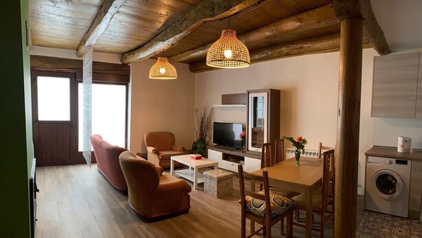 Ein gemütliches Wohnzimmer mit Sofa, Couchtisch und Fernseher. Ein Esstisch mit Stühlen und eine Waschmaschine stehen in der Ecke.