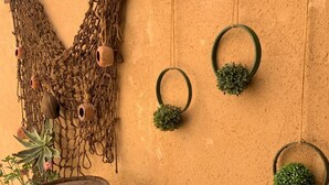 Eine rustikale Wand mit einem Holzbalken, einem Netz aus Holzpfählen und drei hängenden Pflanzgefäßen mit grünen Pflanzen.