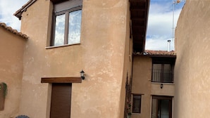 Ein Gebäude mit hellbeige Fassade, einer Holztür und einem Fenster mit Jalousien. Vor dem Haus befindet sich eine kleine Außenbereich-Sitzgelegenheit mit zwei Stühlen und einem Tisch.