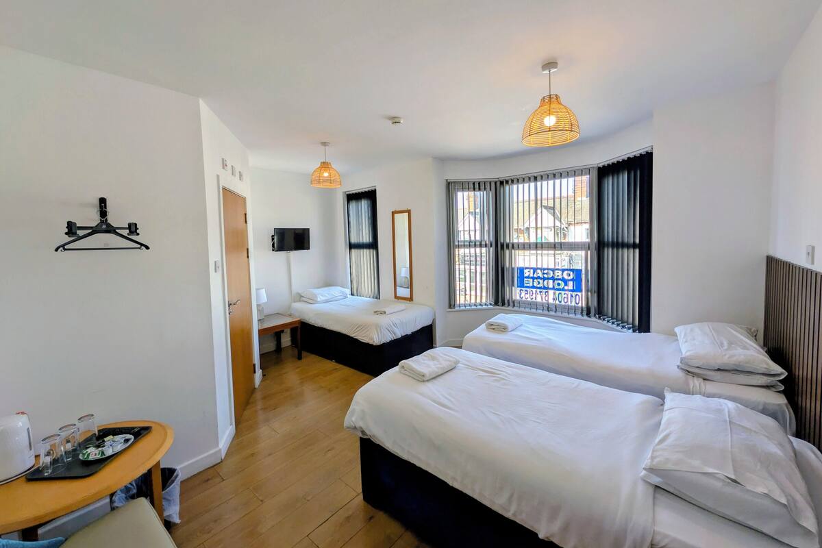 A hotel room with two beds, a TV, a window with blinds, a hanging light fixture, and a small table with a kettle and glasses.