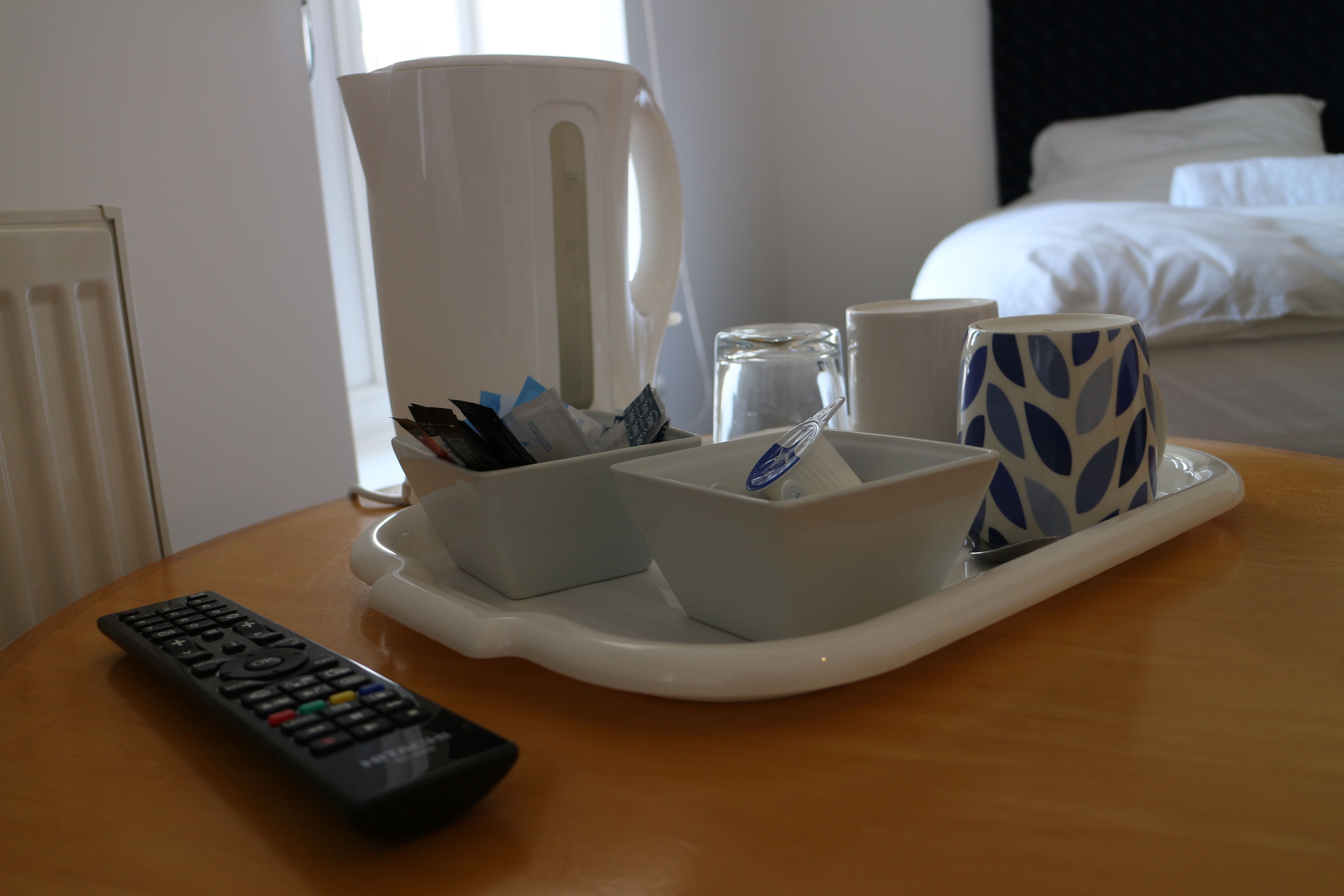 A tray with a remote control, a white pitcher, a glass, and two patterned cups on a wooden table in a hotel room.