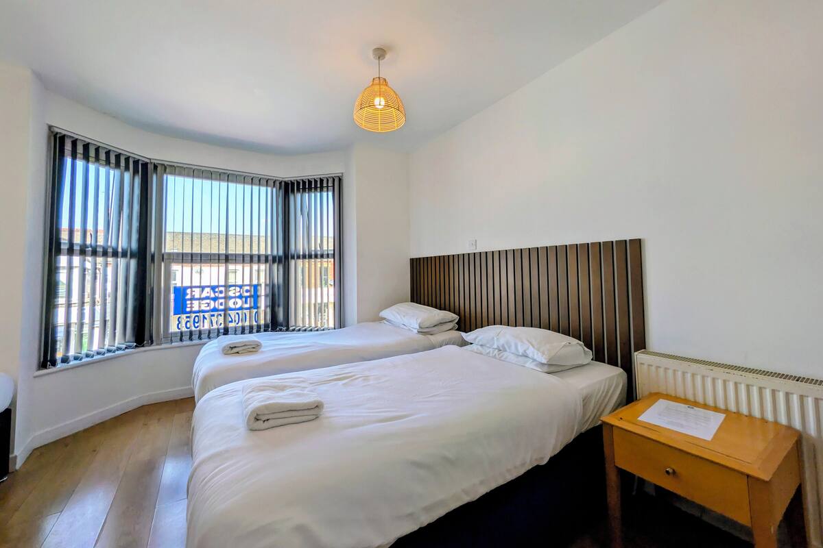 A hotel room with two beds, a wooden nightstand, a radiator, and a window with blinds.