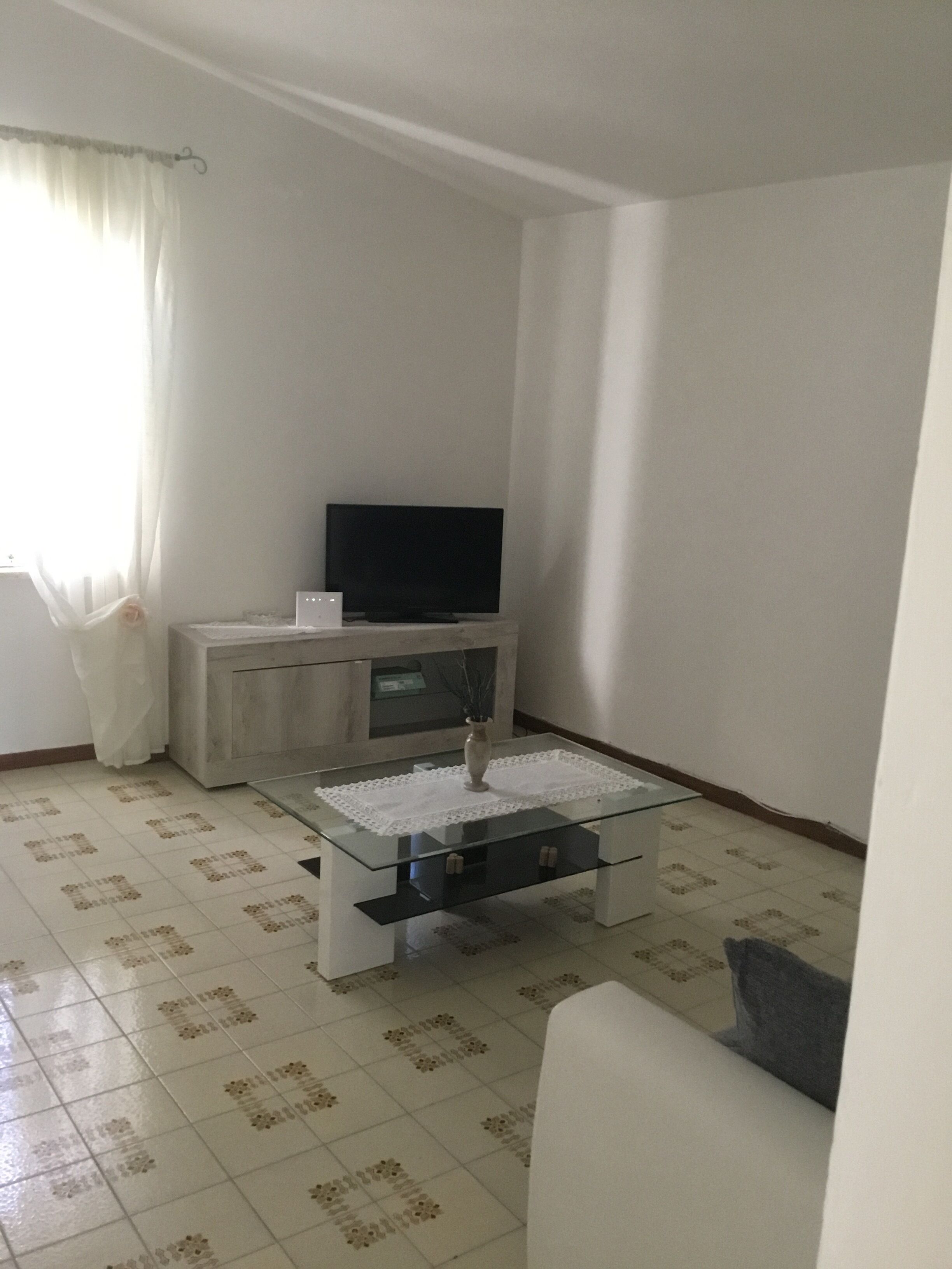 A living room with a glass coffee table, a flat-screen TV on a stand, a white sofa, and patterned floor tiles.