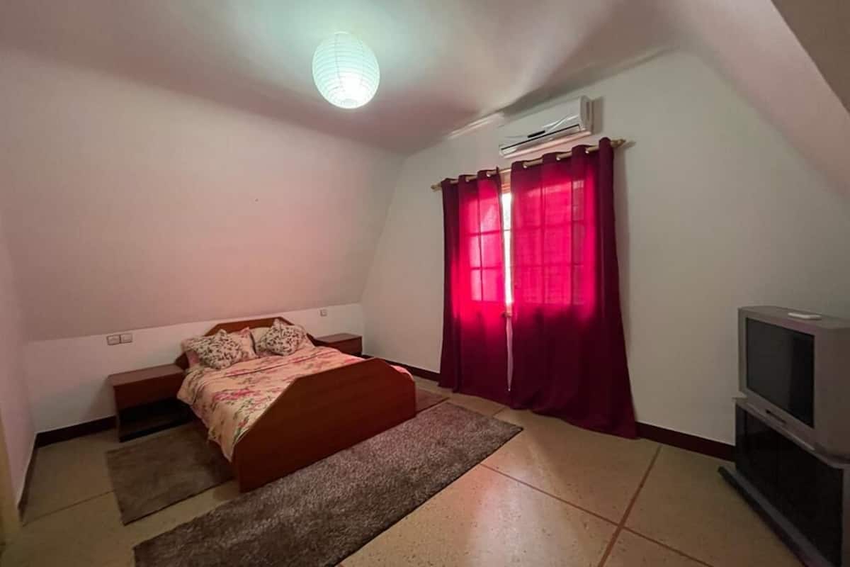 Un dormitorio con cama, televisión, ventana con cortinas y equipo de aire acondicionado.