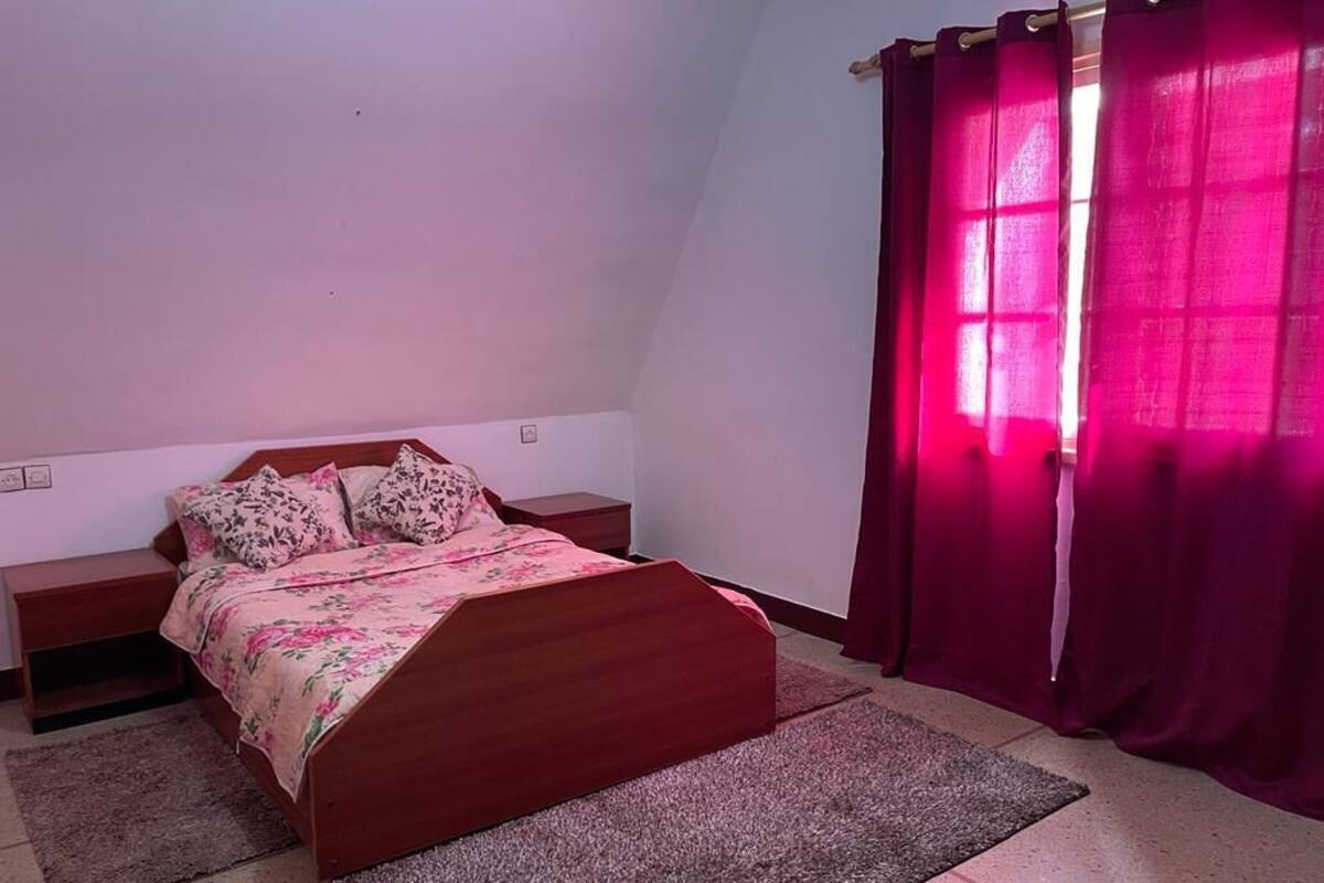 Un dormitorio con una cama de madera, una mesita de noche, una ventana con cortinas rojas y una alfombra pequeña en el suelo.