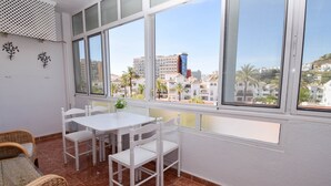 Un balcón con una mesa y sillas blancas, un sof á de mimbre y vistas a edificios y palmeras.